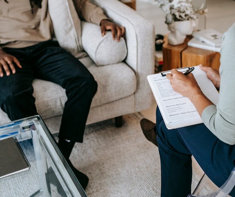 From above crop African American male in casual clothes sitting on comfy couch and answering professional female consultant questions in modern psychotherapy office