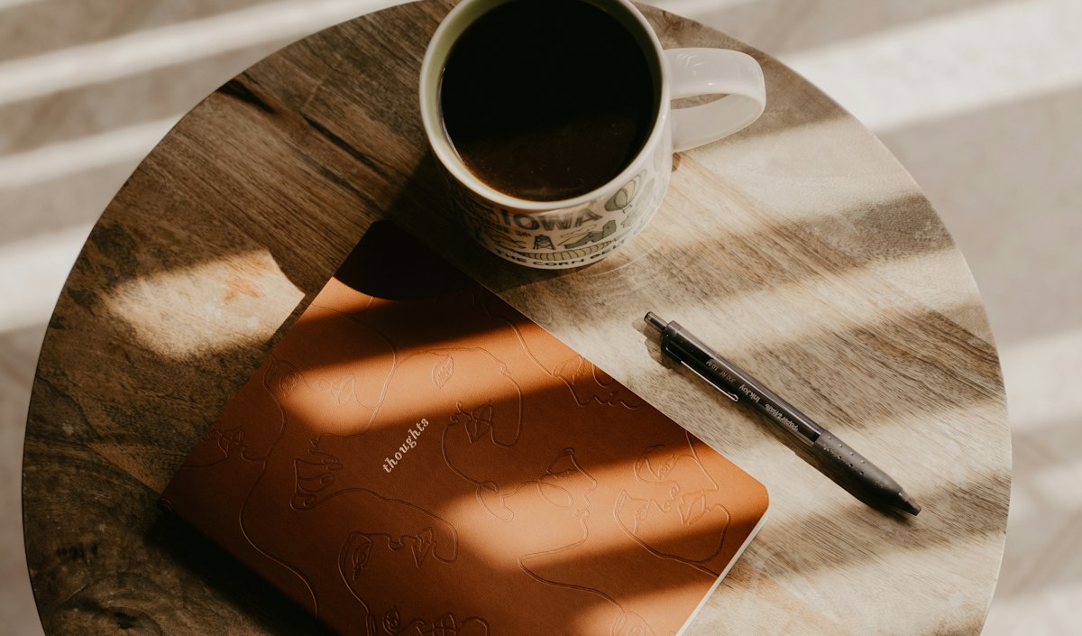 A cup of coffee beside a journal and pen on a wooden table in warm morning light